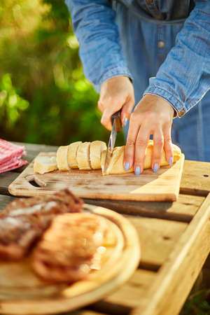 Unrecognizable woman cutting bread for bruschetta on wooden board during weekend barbecue in yard, outdoor, prepare for grilling, summer family picnic, food on the nature.の写真素材