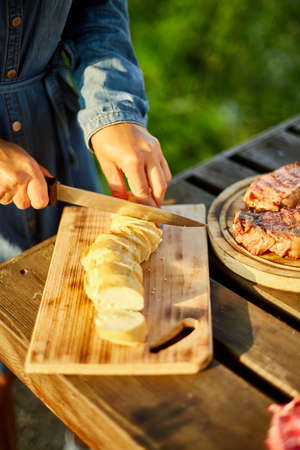 Unrecognizable woman cutting bread for bruschetta on wooden board during weekend barbecue in yard, outdoor, prepare for grilling, summer family picnic, food on the nature.の写真素材