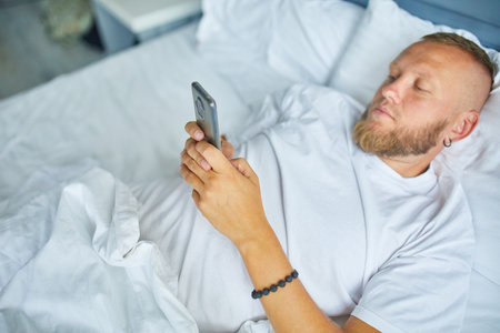 Bearded man using smartphone in morning in cozy while lying on bed white linens at home, male chatting, online surfing internet spend time in bedroomの写真素材