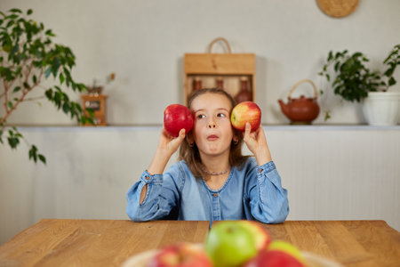 Happy emotional little girl holding red an apple in hands covering eyes at home, smiling children loving fruit healthy snack, vegetarian nutrition for kids, vitamins for children.の写真素材