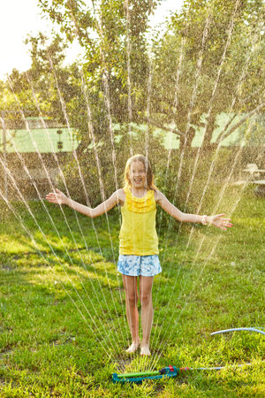 Happy kid girl playing with garden sprinkler run and jump, summer outdoor water fun in the backyard at home, splash on sunny day.の写真素材