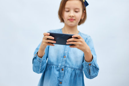 Sad kid girl in casual denim dress use with smartphone, enjoys mobile app, internet addiction, play app on mobile cell phone hold gadget isolated on white background, studioの写真素材