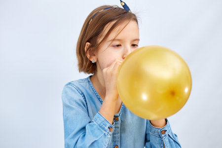 Pretty little girl in casual denim dress blowing, inflate yellow balloon on white background, studioの写真素材
