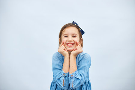 Smiling little girl make trend mini heart sign with fingers in casual denim dress isolated on white...