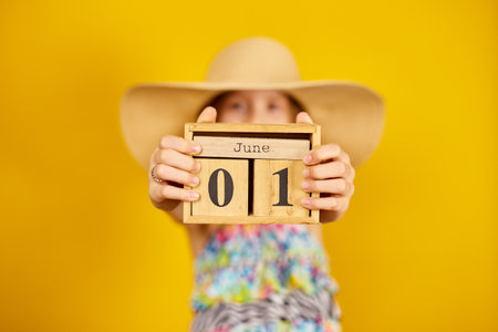 Child teenager girl in swimsuit and straw hat hold in hand wooden calendar on 01 June in studio on yellow background, summer start, summer mood, World Children's Dayの写真素材