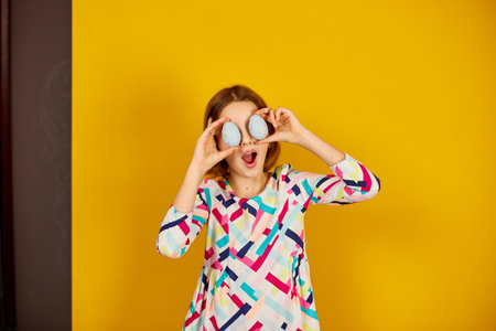 Happy playful teenager girl holding painted Easter eggs covering eyes, on a bright yellow studio...