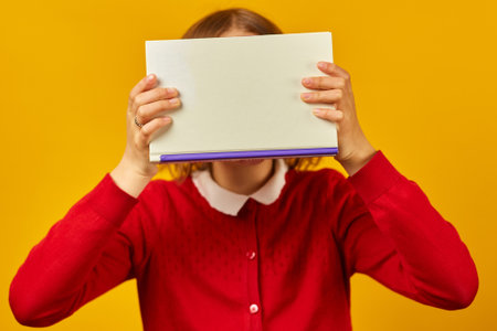 Portrait of happy teenage schoolgirl cover her face by book on yellow studio background, educatin...