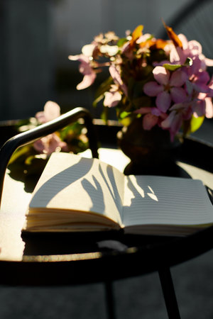 Open notebook and flowers and on the small black table on terrace at home in a sunny day, hardlight, outdoor workspace, summer relaxation, notes, ideas, planningの写真素材