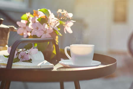 Cup of coffee with steam flowers and notebook on the small black table on terrace at home in a sunny...