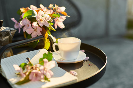 Cup of coffee with steam flowers and notebook on the small black table on terrace at home in a sunny day, outdoor workspace, summer relaxation, coffee break.の写真素材