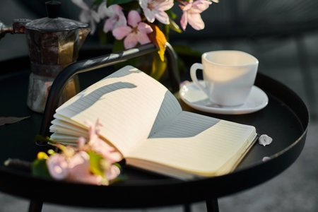 Cup of coffee flowers and notebook on the small black table on terrace at home in a sunny day,...