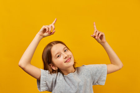 Cheerful, positive ten years teen girl pointing fingers up on workspace area isolated on yellow...