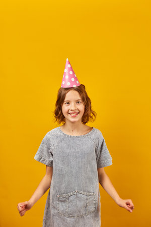 Cheerful, positive ten years birthday girl in party hat, happy, on yellow studio background.