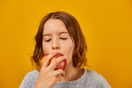 Pretty teen girl, child eat, bites a fresh red apple isolated on studio yellow background, healthy...