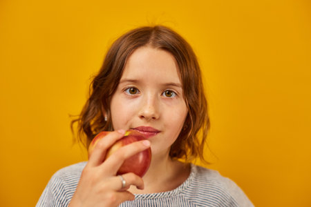 Pretty teen girl, child eat, bites a fresh red apple isolated on studio yellow background, healthy...