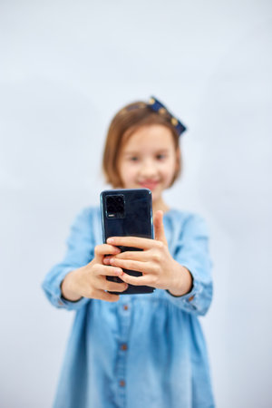 Smiling little girl in casual denim dress hold smartphone do selfie shot on mobile cell phone isolated on white background, studio. lifestyle concept.の写真素材
