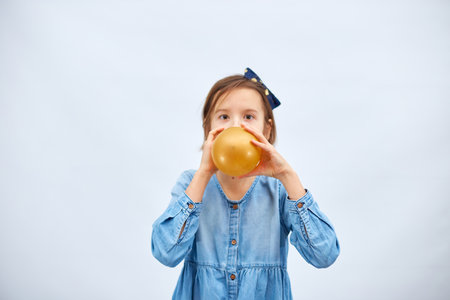 Pretty little girl in casual denim dress blowing, inflate yellow balloon on white background, studioの写真素材