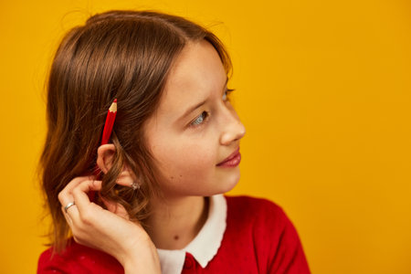Cute teen school girl is thoughtful with red pencil on her ear on yellow studio background, back to school concept, have an idea, copy spaceの写真素材