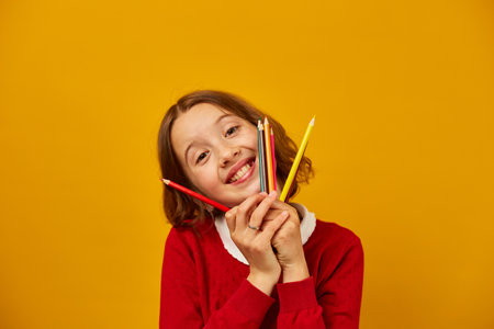 Portrait teen school girl covering her face with colored pencils which holding in hand on yellow studio background, back to school concept, study and writing.の写真素材