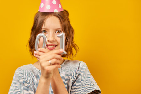Cheerful, positive ten years birthday girl in party hat with candles, happy, on yellow studio...