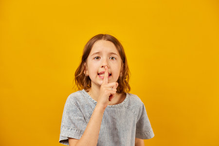 A young girl stands before a vivid yellow backdrop, placing her index finger over her lips in a...