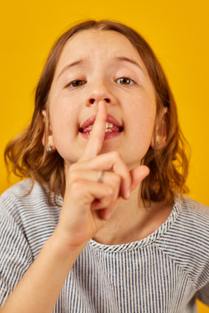 A young girl stands before a vivid yellow backdrop, placing her index finger over her lips in a...