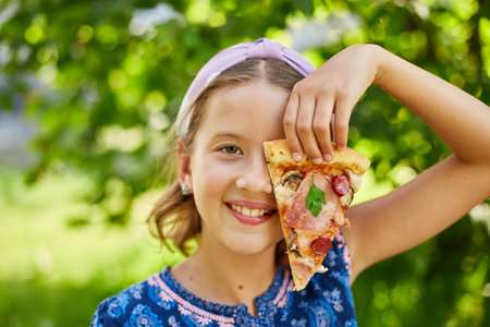 Cheerful Young Girl Enjoying a Slice of Pepperoni Pizza Outdoors on a Sunny Dayの写真素材