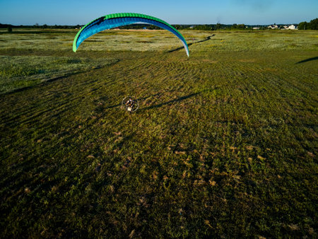 A single paraglider enjoys an exhilarating flight over expansive green fields under the clear blue...