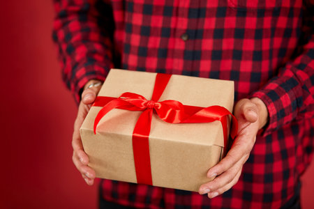 Close-up of hands holding a gift box with a red ribbon against a red background.の写真素材