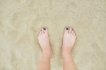 A close-up view of a persons bare feet with dark blue toenail polish on sandy beach texture.