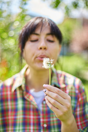 A woman gently blows on a dandelion, scattering seeds in a vibrant garden, Springtime