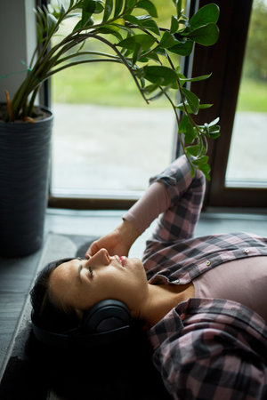 Lying on a mat near an open door, a woman enjoys music with her headphones, a plant beside her.