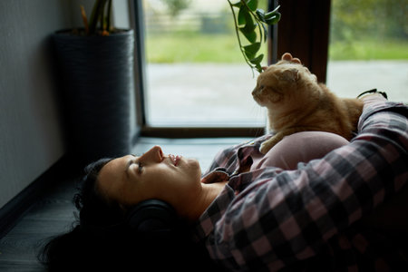 A woman lies back with headphones, enjoying the company of her cat near a plant.の写真素材