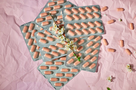 Blister packs with capsules scattered on a textured pink background, alongside small white flowers.