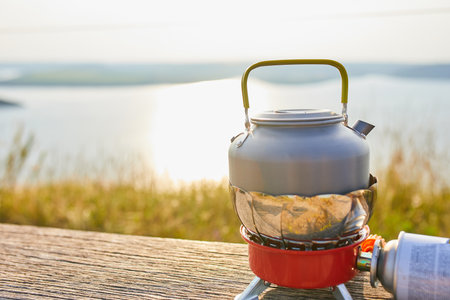 A kettle is placed on a portable stove near a serene lake while the sun sets in the background. The warm glow enhances the calm atmosphere, ideal for outdoor cooking.の写真素材