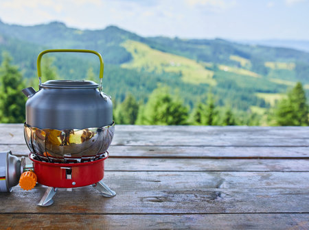 A kettle sits on a red camping stove, surrounded by a breathtaking view of green mountains and trees, capturing a peaceful outdoor moment in nature.の写真素材