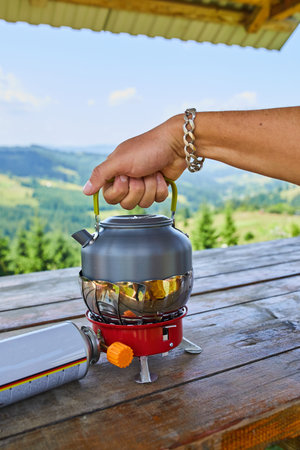 A hand lifts the lid of a metallic kettle sitting on a portable stove. The backdrop features stunning mountains under a clear blue sky, suggesting a peaceful outdoor experience.の写真素材