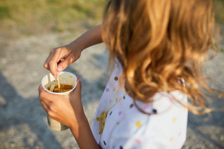 A young child is savoring a warm snack from a cup while standing in a relaxed outdoor setting, surrounded by natural beauty and cheerful atmosphere.の写真素材