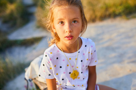 A young girl sits on the beach, wearing a colorful polka dot shirt and a necklace. The sun sets in the background, casting a warm glow on her face as she gazes into the camera.の写真素材
