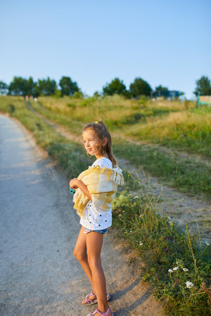 A young girl stands by a winding path, wearing shorts and a yellow shawl. She enjoys a sunny day surrounded by green grass and a clear blue sky, smiling gracefully.の写真素材