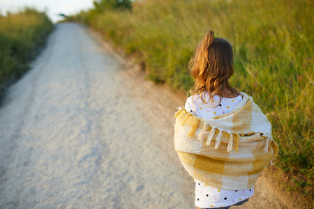 A young girl strolls along a dirt path, enjoying the summer sun. She wears a light shawl over her shoulders and denim shorts, surrounded by green hills and nature.の写真素材