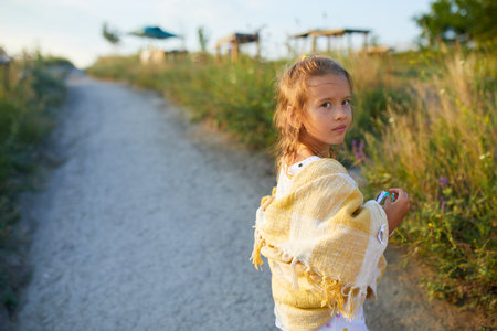 A young girl stands by a winding path, wearing shorts and a yellow shawl. She enjoys a sunny day surrounded by green grass and a clear blue sky, smiling gracefully.の写真素材
