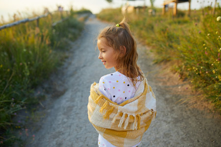 A young girl stands by a winding path, wearing shorts and a yellow shawl. She enjoys a sunny day surrounded by green grass and a clear blue sky, smiling gracefully.の写真素材