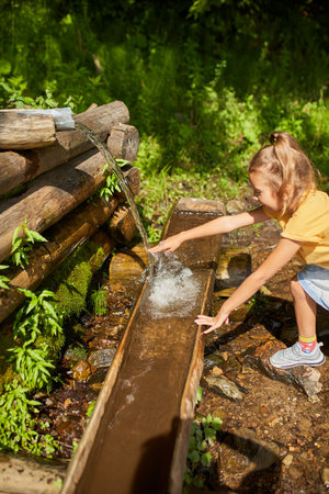 A young girl stands by a natural spring, splashing water on her face to cool off on a warm sunny day. Surrounded by lush greenery, she enjoys a delightful moment in nature.の写真素材