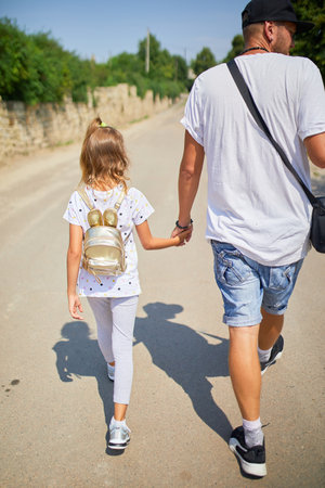 A father walks alongside his daughter on a sunny day, holding hands as they explore a tranquil road. The little girl carries a small backpack and smiles brightly, sharing a joyful moment.の写真素材