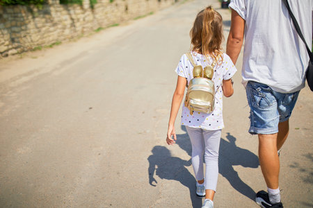 A father walks alongside his daughter on a sunny day, holding hands as they explore a tranquil road. The little girl carries a small backpack and smiles brightly, sharing a joyful moment.の写真素材