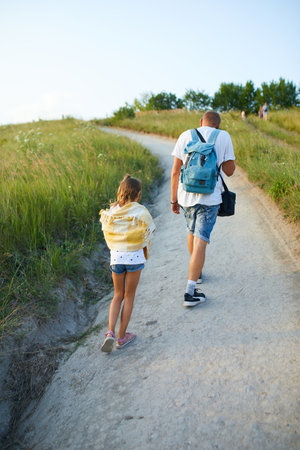 A father and his young daughter stroll along a winding path surrounded by tall grass on a sunny day. The warm light creates a serene atmosphere as they enjoy their time together.の写真素材
