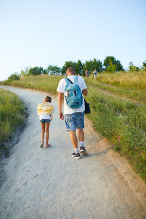 A father and his young daughter stroll along a winding path surrounded by tall grass on a sunny day. The warm light creates a serene atmosphere as they enjoy their time together.の写真素材