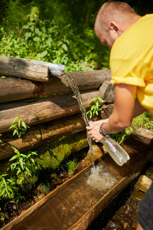 A traveler fills a clear bottle with fresh water from a wooden spring while surrounded by vibrant greenery on a sunny summer day. The serene atmosphere enhances the adventurous spirit.の写真素材