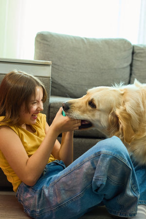 A cheerful teen girl sits on the floor, laughing as she holds a colorful ball towards a golden retriever. They are in a cozy living room filled with natural light.の写真素材